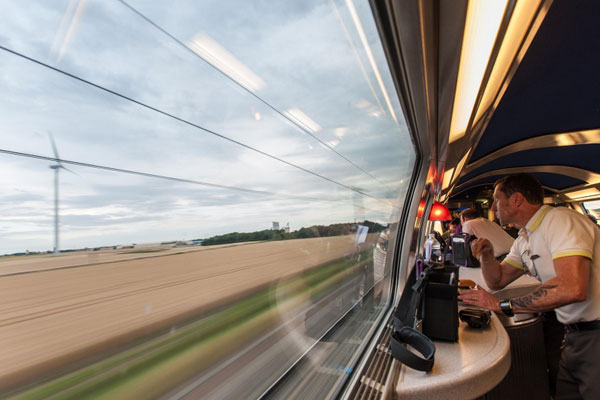 A man is looking out the window of a TGV that is passing cornfields and windmills.
