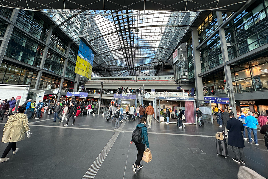 The main level of the Berlin Central station is crowded.  A train is loading passengers on the upper level.