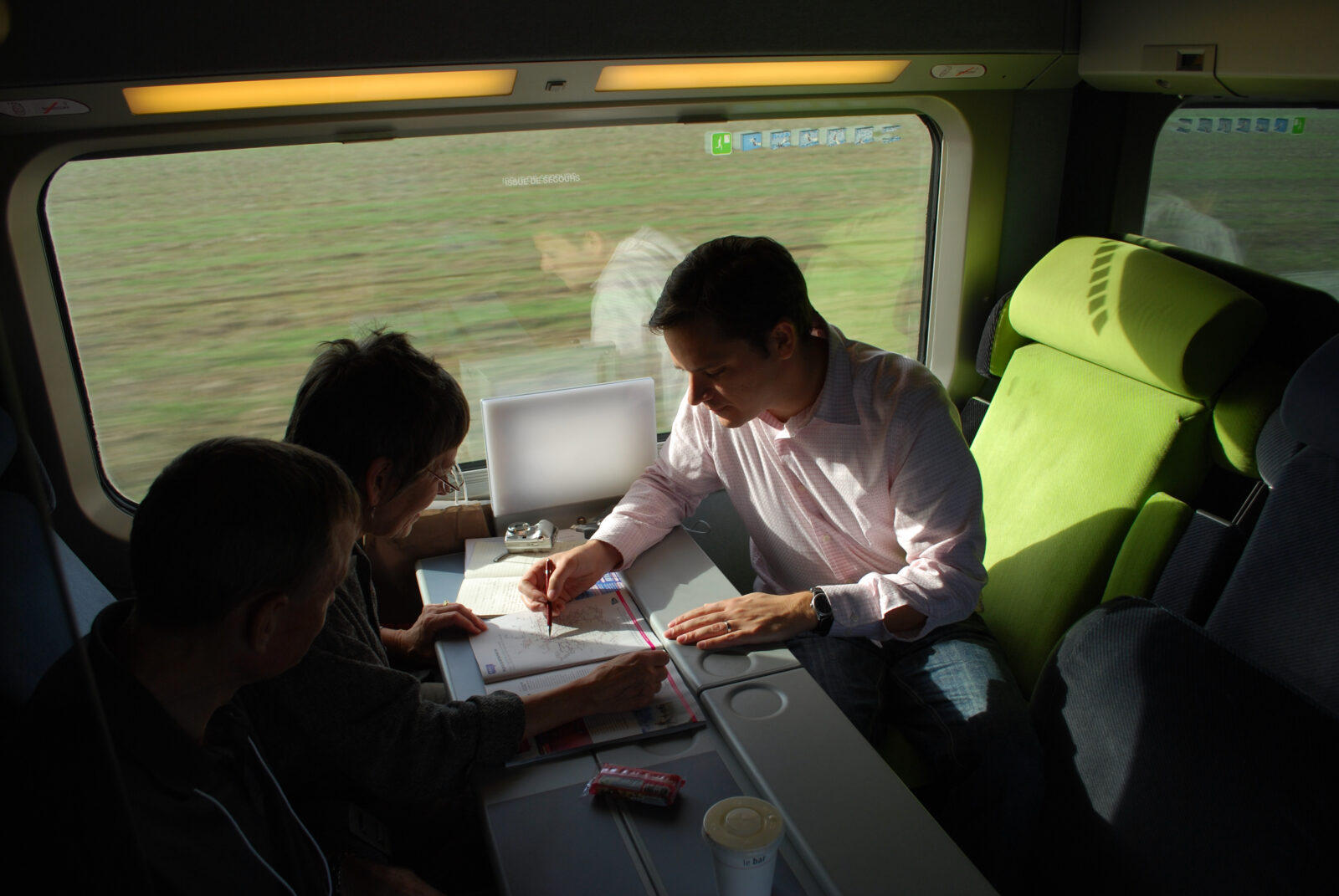 A man pointing at a document while sitting across from two freinds at a table in a high-speed train with field blurbed in background.