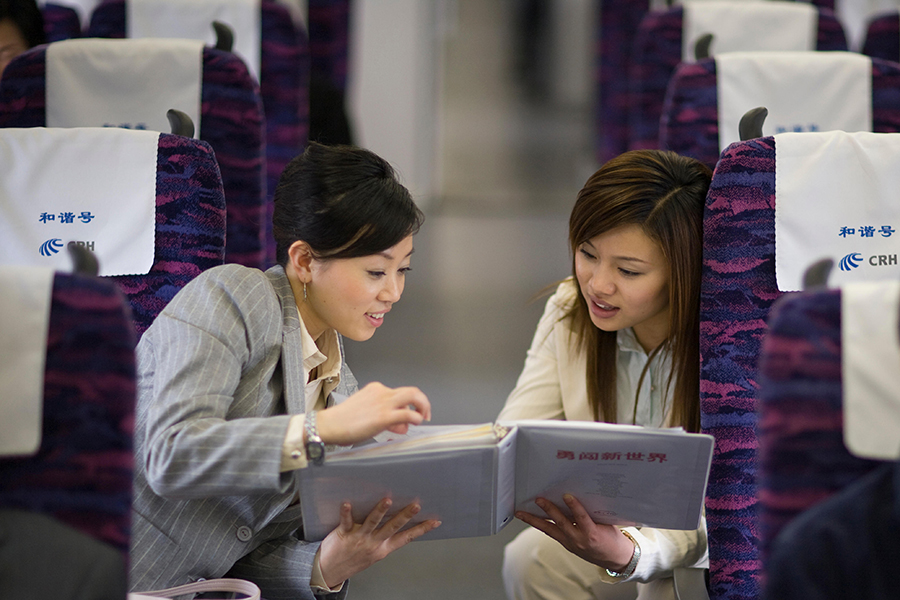Two woman talking across the aisle of a high-speed train while looking at a 3-ring binder.