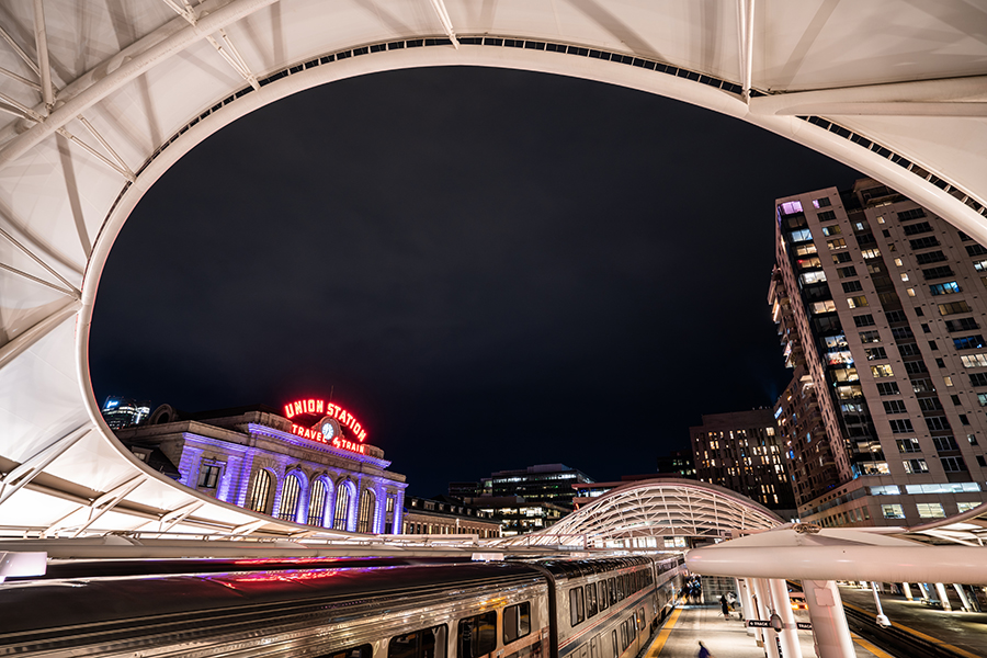 An Amtrak train is in the station at Denver Union Station at night. Passengers are on the platform. The iconic neon Union Station Travel by Train sign is lit up.