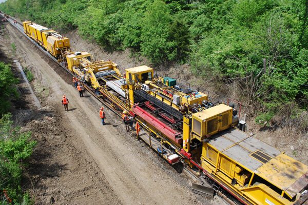 A crew is replacing track with a machine that the front rides on the old track and the back rides on the new track. The middle replaces the track and ties all at once.