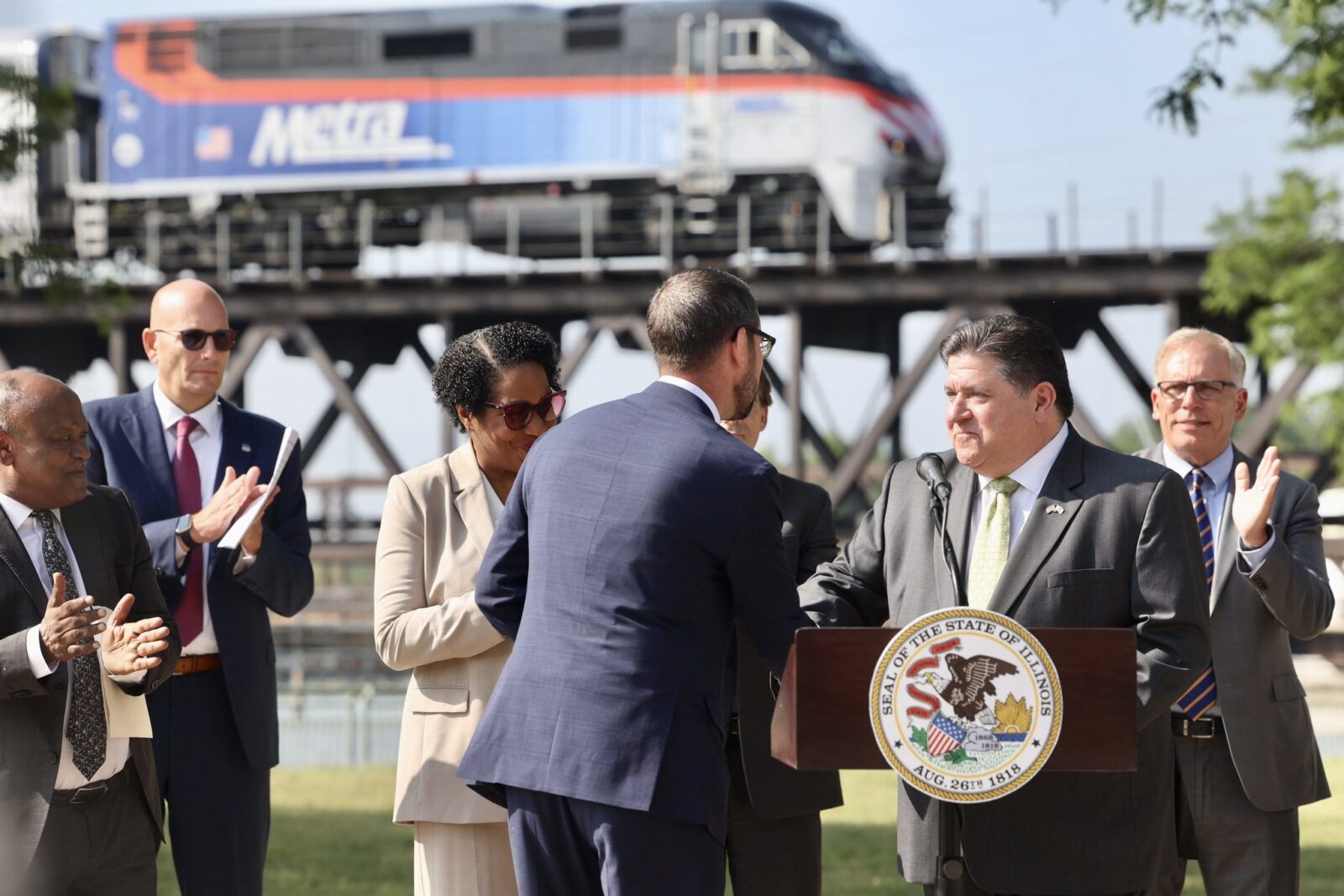Gov. Pritzker announces that Metra will serve Rockford at a press conference with a Metra locomotive visible on a bridge in the background.