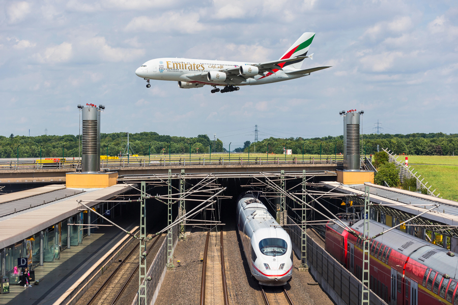 An A380 plane is landing at Dusseldorf airport. Railroad tracks run under the runway and the station is in the foreground. A high-speed train is passing through the station and a local train is stopped at he station.