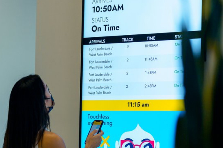 A woman is looking at the departures board at a Brightline station. it shows hourly departures.