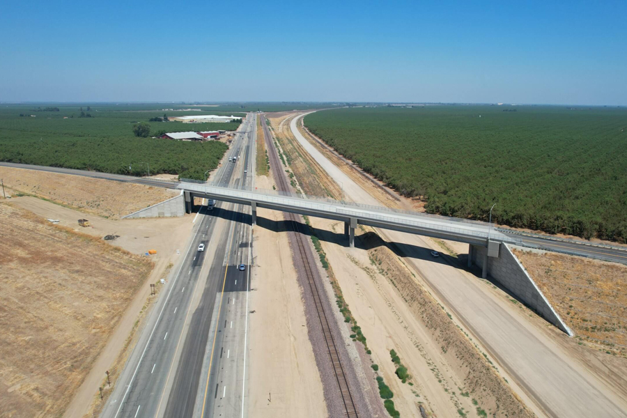 A overhead view of a new highway bridge over a four-lane highway, a railroad and the new right-of-way for the California high-speed line.