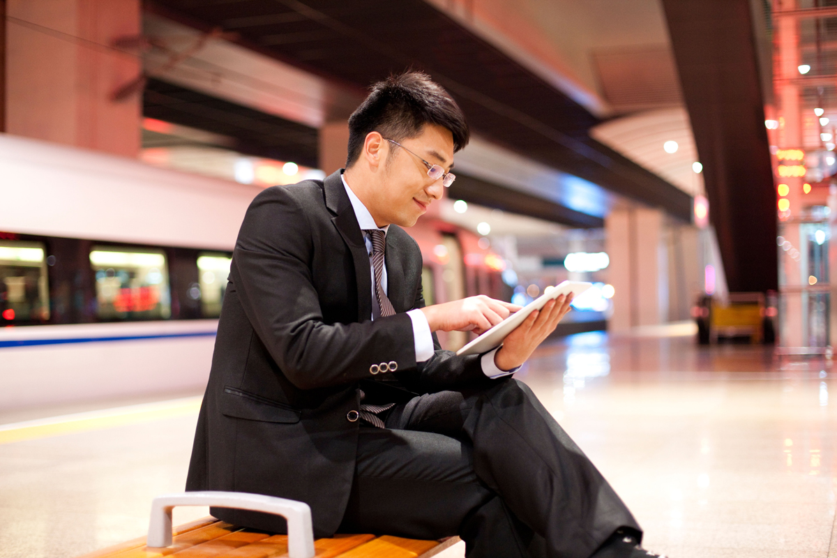 A dude is using an IPad on a station platform wth a high-speed train in the background.