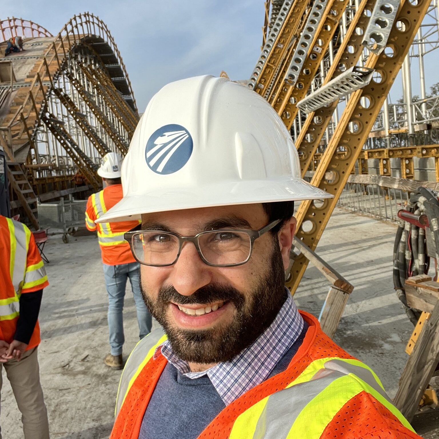 A man in a construction vest and hard helmet stands before a viaduct under construction