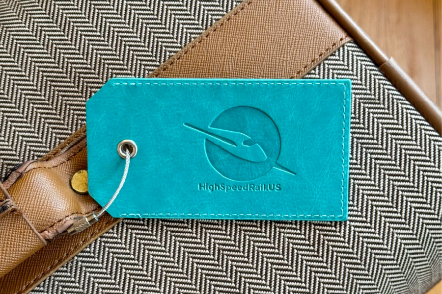 A teal High Speed Rail Alliance luggage tag attached to a tan herringbone suitcase by a metal wire.